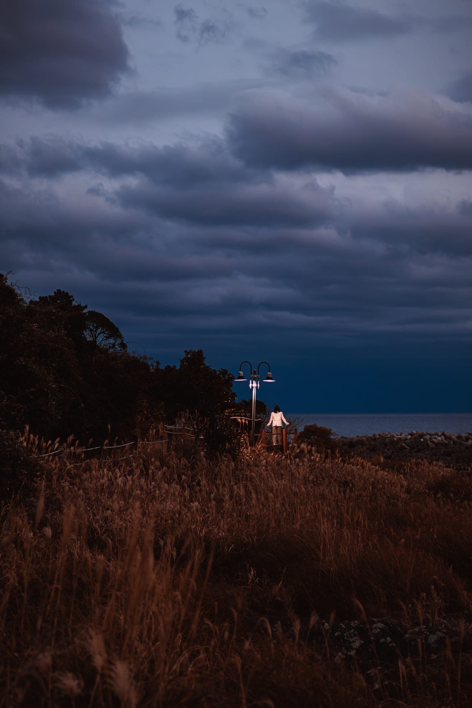 A rocky shore at dusk under heavy clouds, one small light by the sea