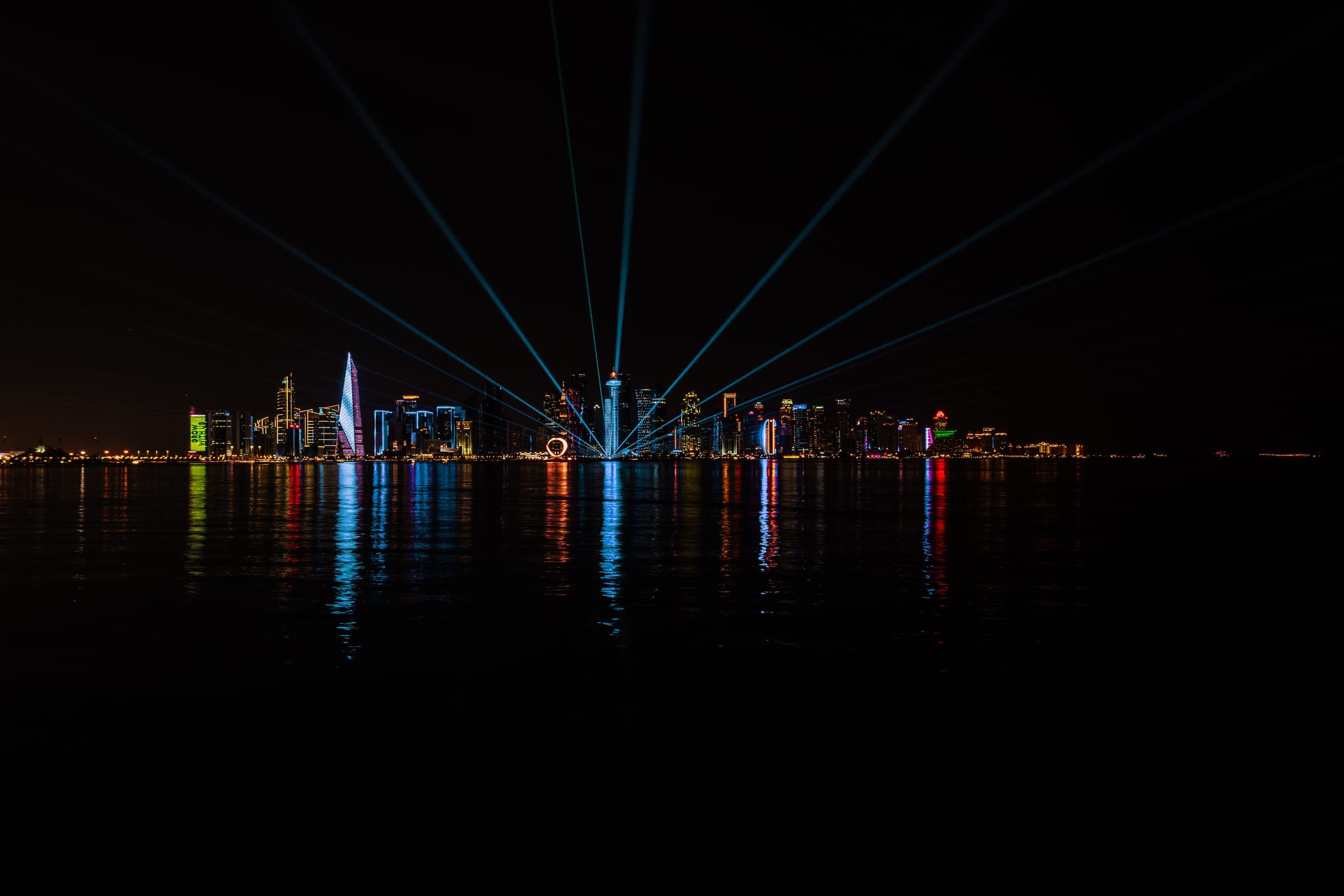 A city skyline at night across the water, beams of light fanning into the sky