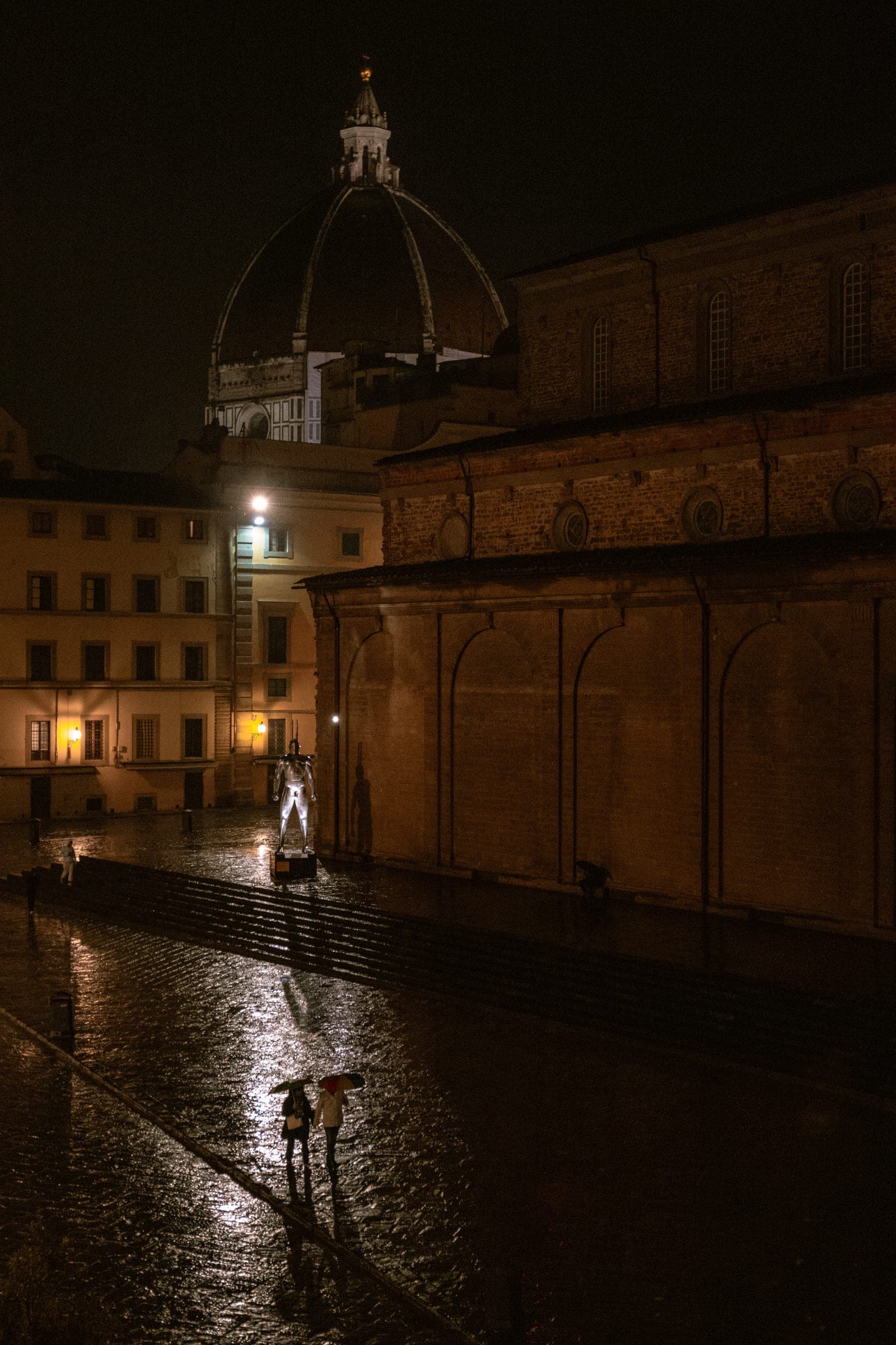 A cathedral dome over a wet street at night, one figure under the lamps