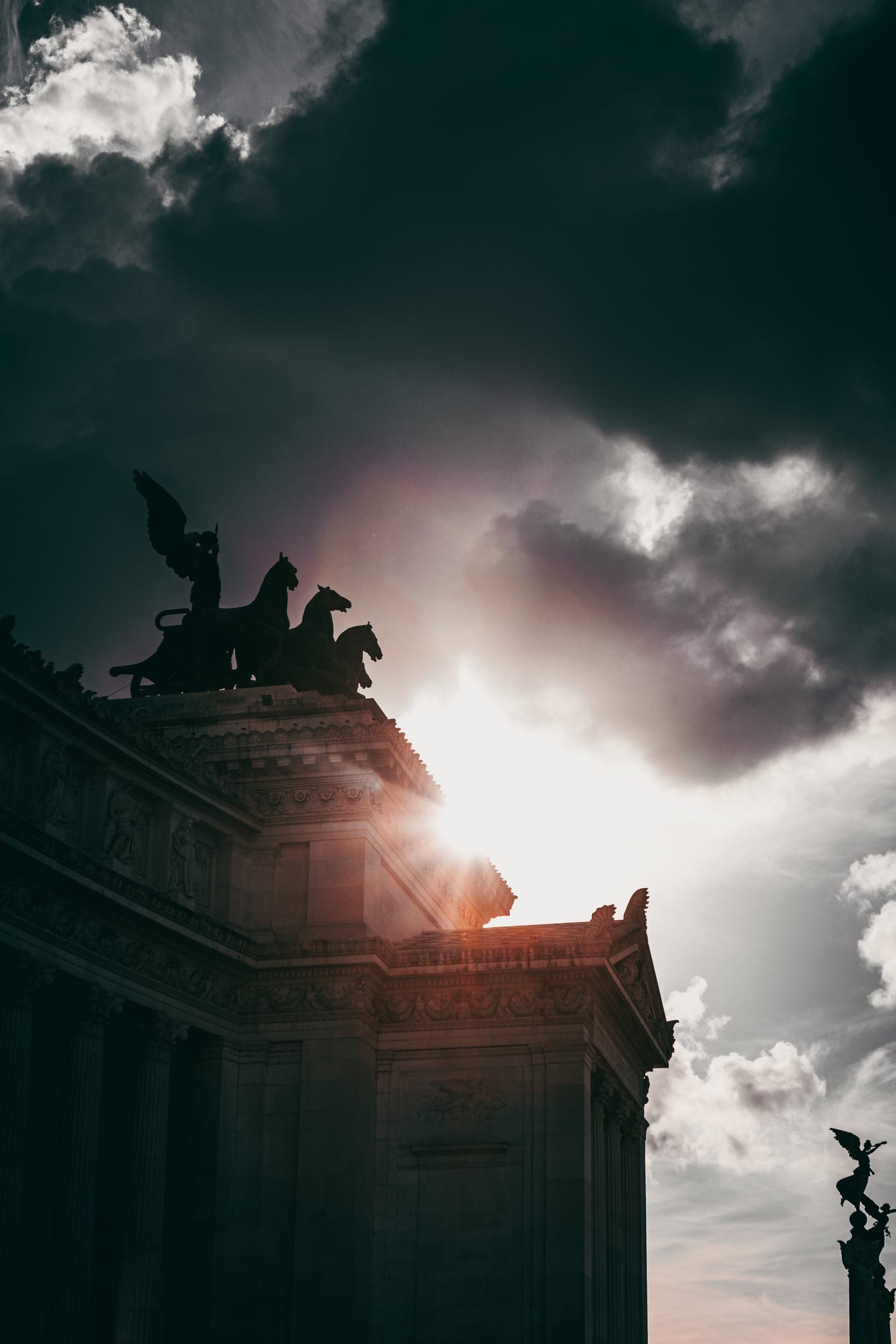 A bronze chariot group on a monument, the sun bursting through dark clouds behind it
