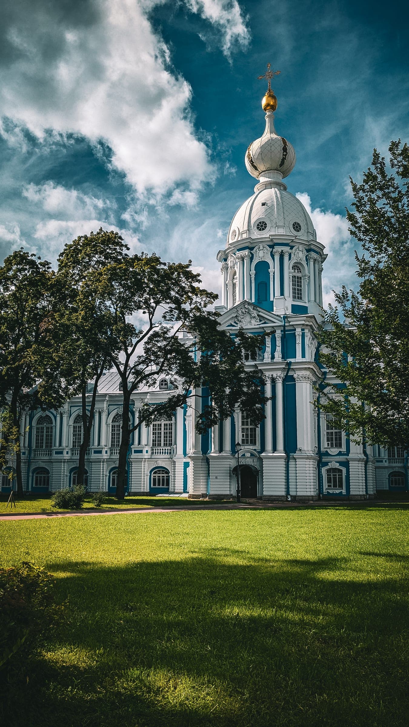 The white and blue baroque Smolny Convent behind trees and a sunlit lawn