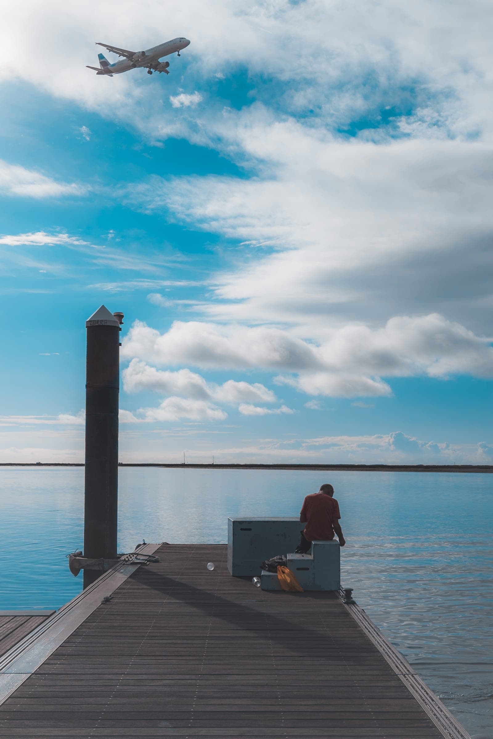 A person sitting at the end of a wooden pier over flat water, a plane crossing the sky